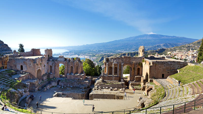 Antikes Theater von Taormina auf Sizilien mit Blick auf die Küste und den dampfenden Vulkan Ätna unter blauem Himmel