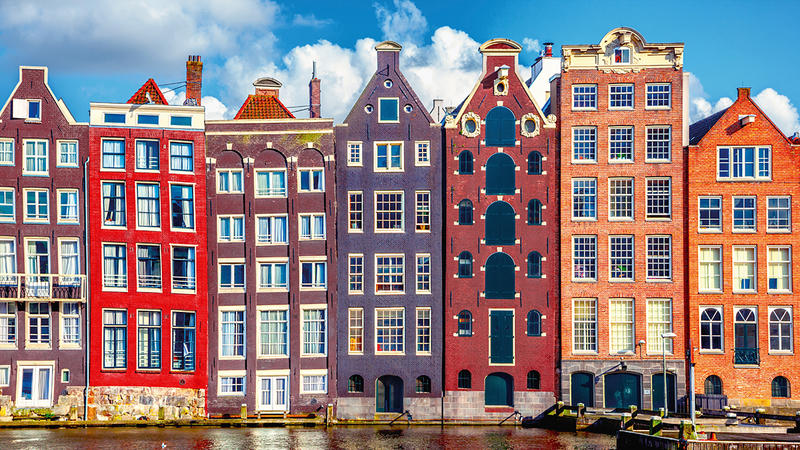 Bunte historische Giebelhäuser direkt am Wasser einer Amsterdamer Gracht bei sonnigem Wetter mit blauem Himmel