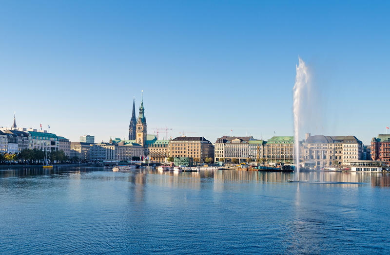 Blick über die Hamburger Binnenalster mit Fontäne auf das Rathaus und die Silhouette der Stadt bei strahlendem Sonnenschein