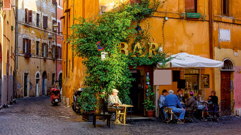 Gäste an Außentischen einer Bar in einer sonnigen, charmanten italienischen Gasse mit ockerfarbenen Häuserfassaden