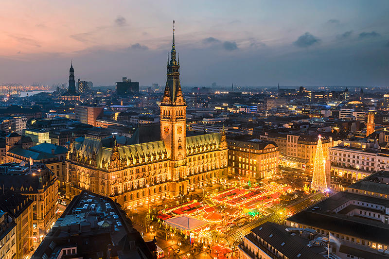Beleuchteter Hamburger Weihnachtsmarkt auf dem Rathausmarkt bei Dämmerung mit Blick über die abendliche Stadtlandschaft
