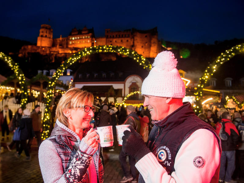 Paar mit Glühwein auf dem weihnachtlichen Heidelberger Marktplatz vor der beleuchteten Schlossruine am Abend